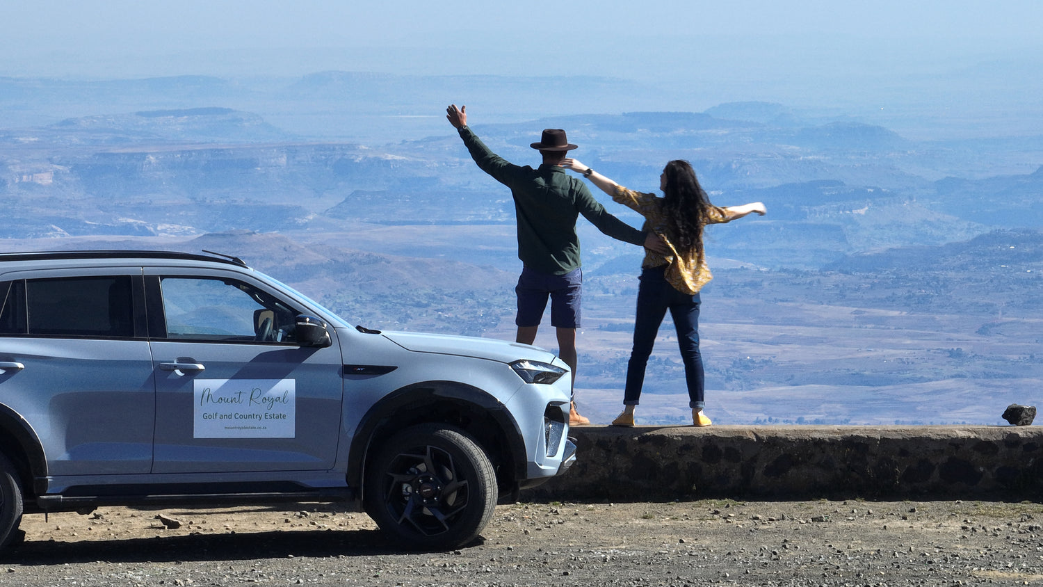 A light blue SUV parked on a rocky overlook with two people standing on a stone wall in front of it, arms outstretched, overlooking a vast mountain landscape under a clear blue sky.