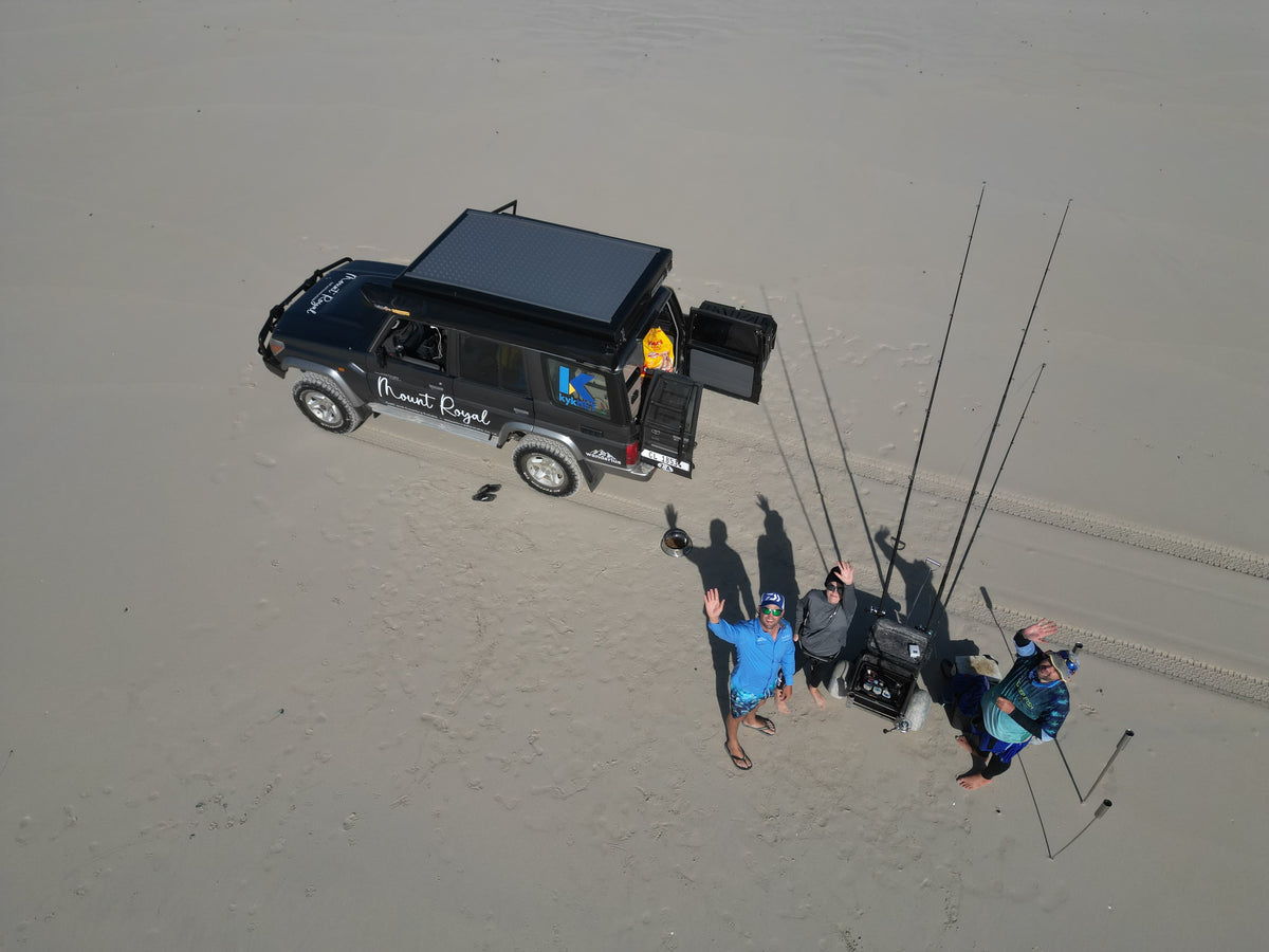 An aerial view of a black custom off-road vehicle with a roof top mounted on a sandy beach, with three people standing next to it holding fishing rods and tackle gear.
